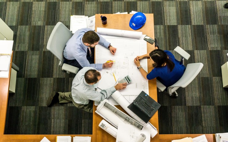 Mesa de escritório vista de cima com dois homens e uma mulher em uma reunião de gestão de obras. Sobre a mesa há uma planta de arquitetura, notebook e um capacete de construção.