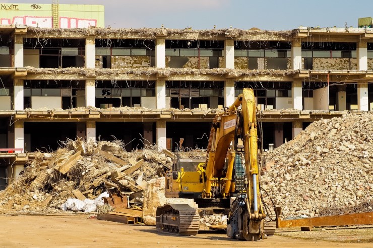 imagem de um prédio de três andares em demolição, na frente estão duas pilhas de entulho e uma escavadeira amarela 